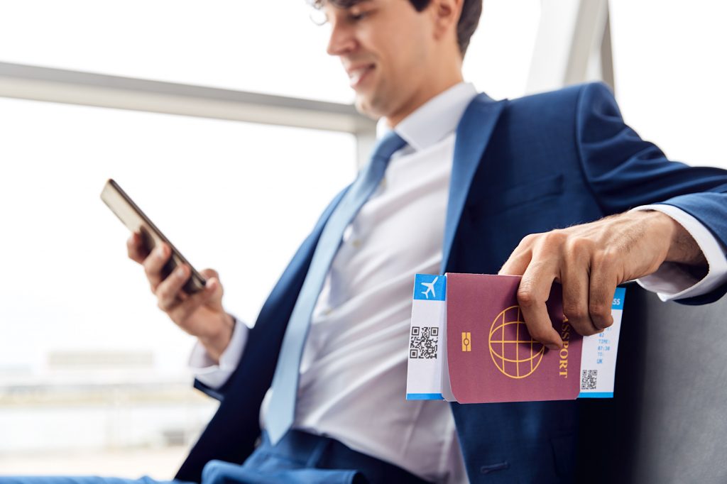 Businessman With Passport And Boarding Pass Sitting In Airport Departure Lounge Using Mobile Phone